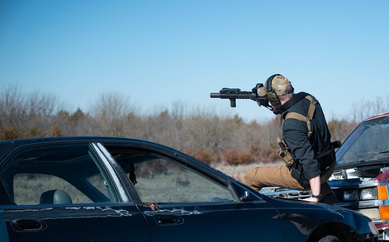 Competitor sliding across a car hood while aiming a rifle on a daytime Run N Gun vehicle stage at The Burial Mound