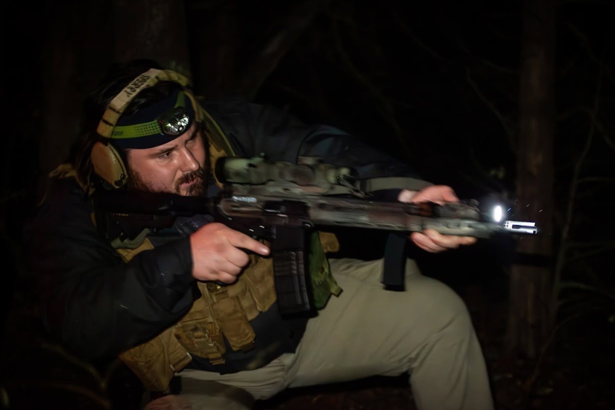 Competitor in a headlamp and plate carrier kneeling with a weapon-lit rifle in the woods during the Twilight Biathlon